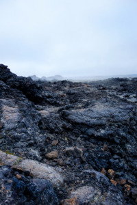 The image depicts a rocky area with a mountain in the background, resembling the surface of the moon. The terrain is characterized by volcanic rock and igneous formations, with a vast expanse of rocky ground and a foggy sky.