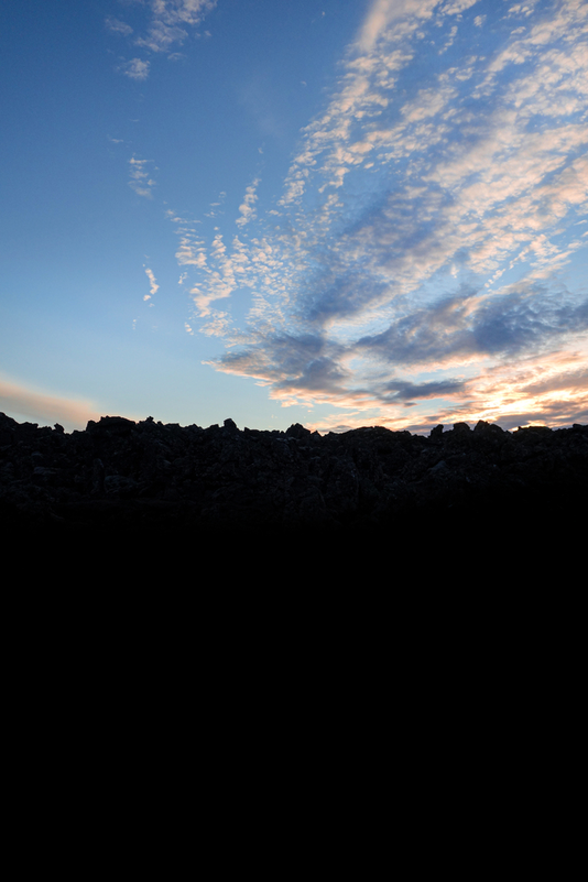 The image depicts a serene sky with clouds, possibly during sunset or sunrise. The horizon is not visible. The rocks on the mountain in the fronts are dark due to the sun cannot reach them anymore. The overall scene is of an outdoor natural landscape.