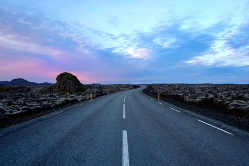The image depicts a road running through a lifeless landscape with a rock formation on the side. The sky in the background is pink and blue, and the surroundings appear to be made of lava.