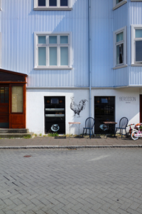 The image shows a building in Reykjavik, Iceland with a table and chairs outside. The scene captures a city street with a parked vehicle and sidewalk.